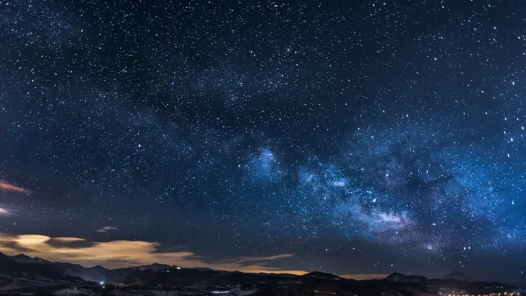 Rozgwieżdżone nocne niebo nad ciemnym zarysem górskiego krajobrazu, tworzące spokojną atmosferę.