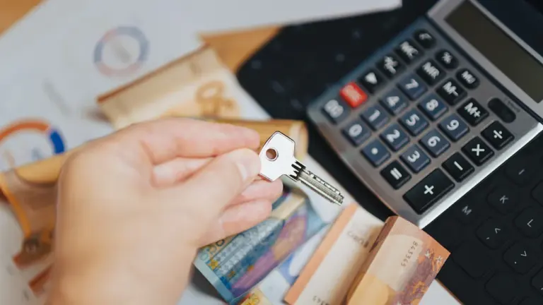 Dłoń trzymająca klucz do domu, kalkulator oraz banknoty na dokumentach finansowych.