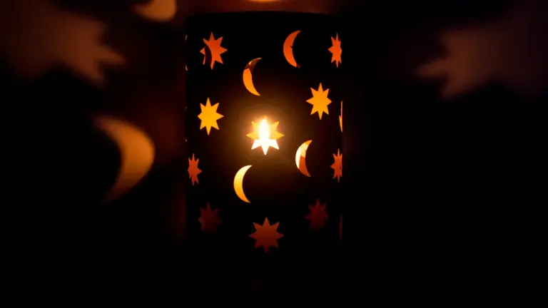 Ciemny lampion z wyciętymi wzorami gwiazd i księżyców emitujący nastrojowe ciepłe światło.